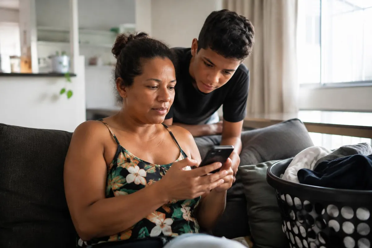 Mãe e filho adolescente no sofá da sala olham juntos para um celular, com uma cesta de roupas ao lado