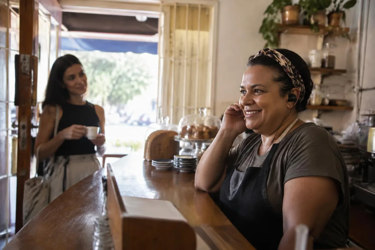 Dona de cafeteria sorri enquanto fala ao fone de ouvido atrás do balcão, com uma cliente aguardando ao fundo